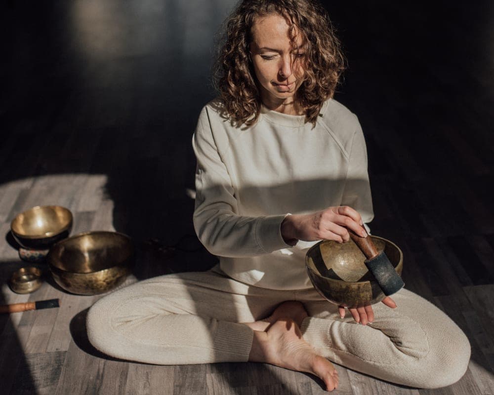 A practitioner cross-legged on a wooden floor with brass singing bowls, mid-practice.