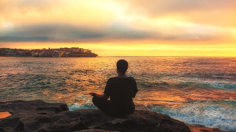 A silhouette seated above the ocean as gold light spreads across the water.