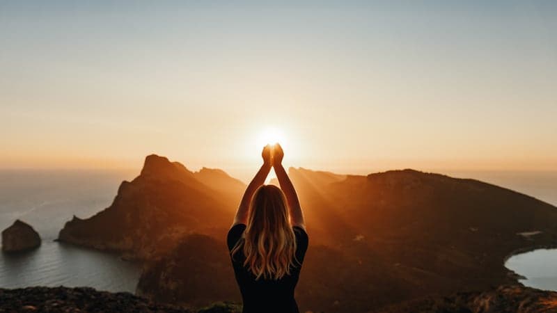 A figure on a mountain ridge raising the setting sun between cupped hands.