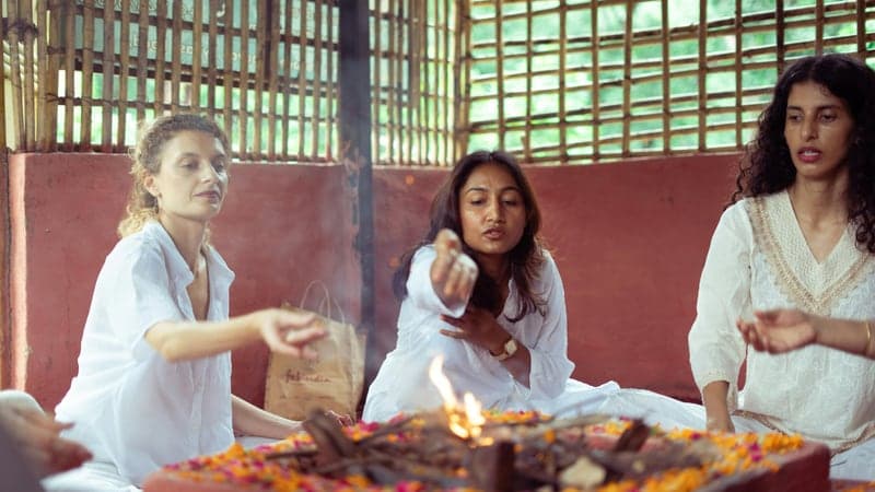 Three women in white at a small fire ceremony, calm and unhurried.